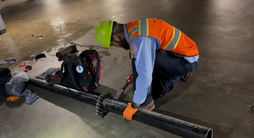 Professional plumbing technician using a pipe-cutting tool on a large black pipe while wearing protective safety equipment.