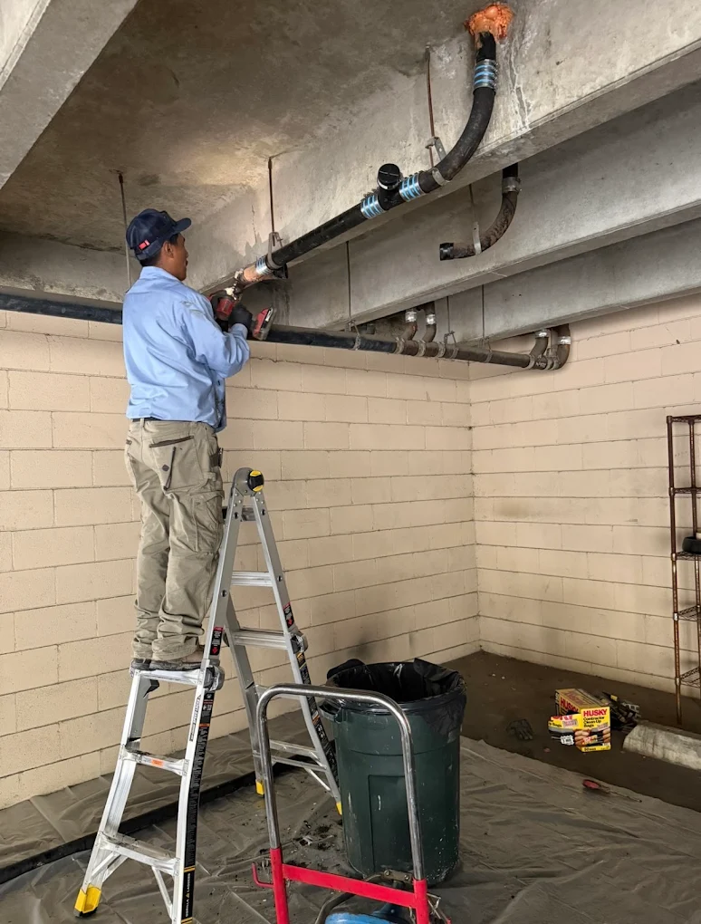 Professional plumber working on overhead drain piping and pipe connections inside a commercial-style building.