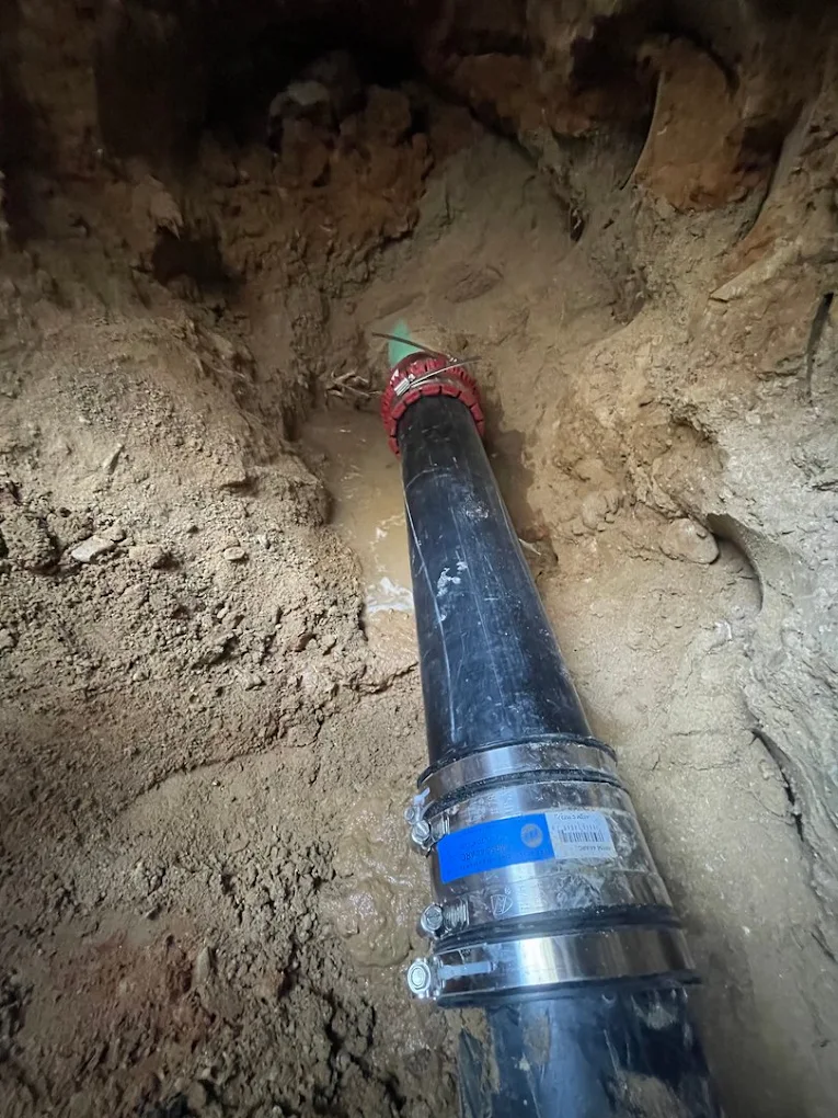 Exposed underground drain pipe with rubber couplings, metal clamps, and standing water around the repaired pipe connection.