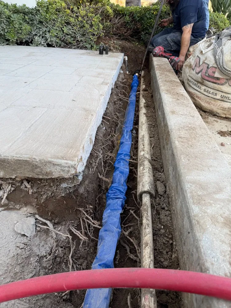 Plumber working on underground pipe installation in a residential trench alongside an existing plumbing line.